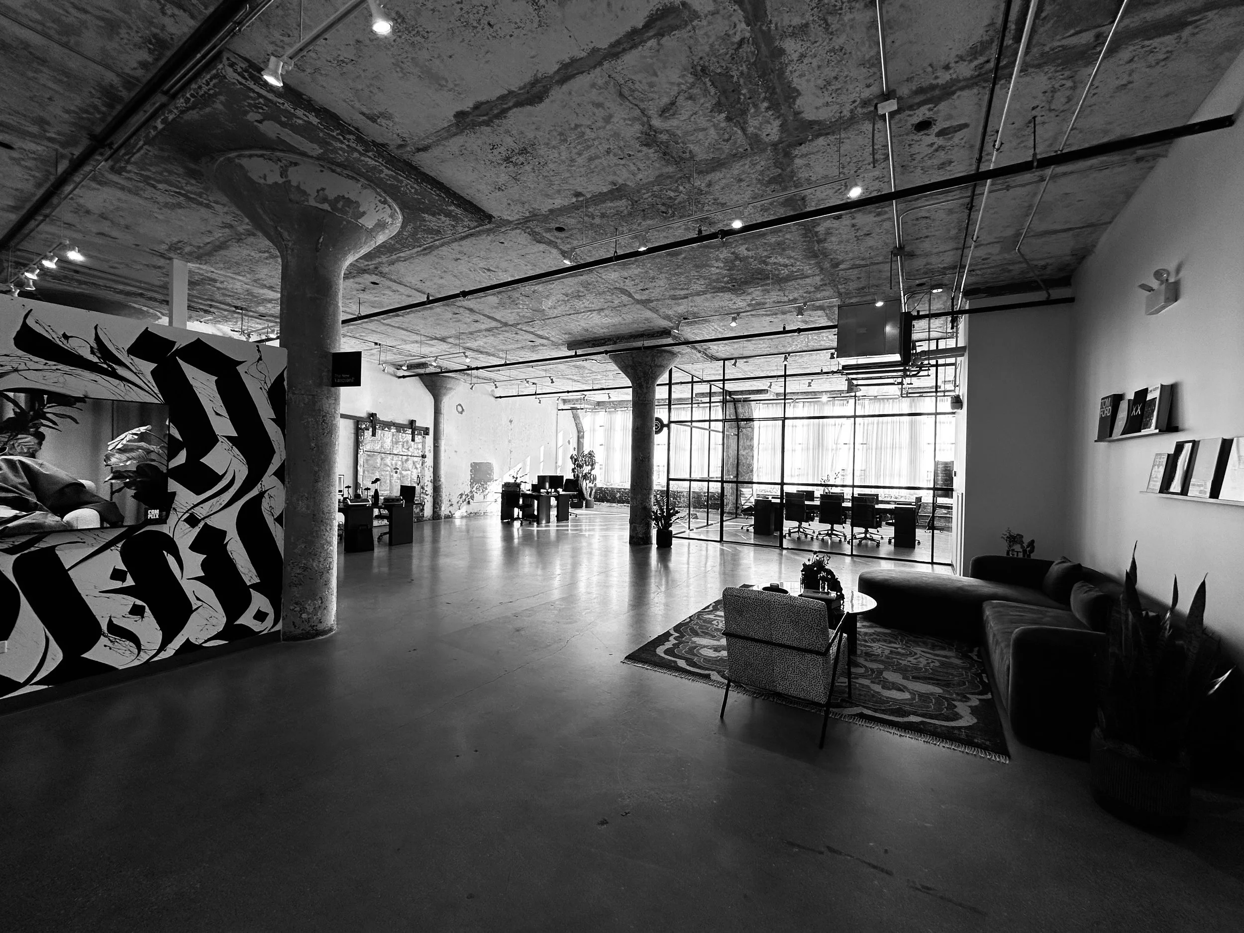 Interior of a modern, industrial-style office space with exposed concrete ceilings, large windows, office desks, seating area with a rug, wall art, and plants.