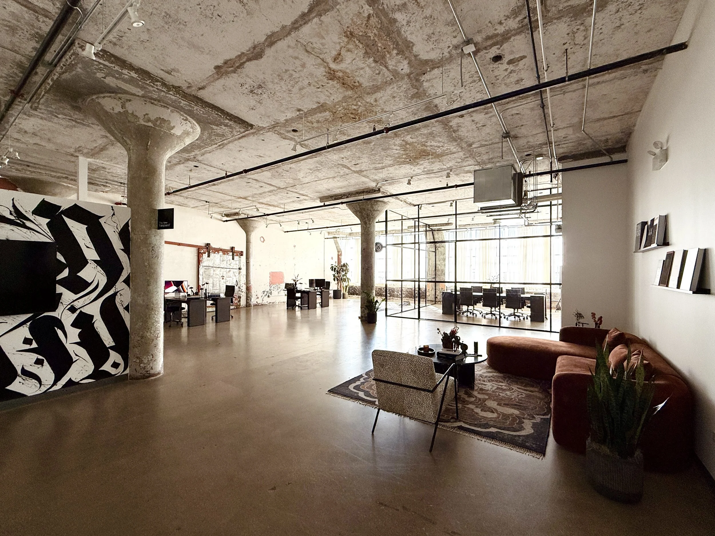 Open-plan loft office with exposed concrete ceiling and large windows. Furnished with workstations, a lounge area with a patterned rug, sofa, armchair, and decorative plants.