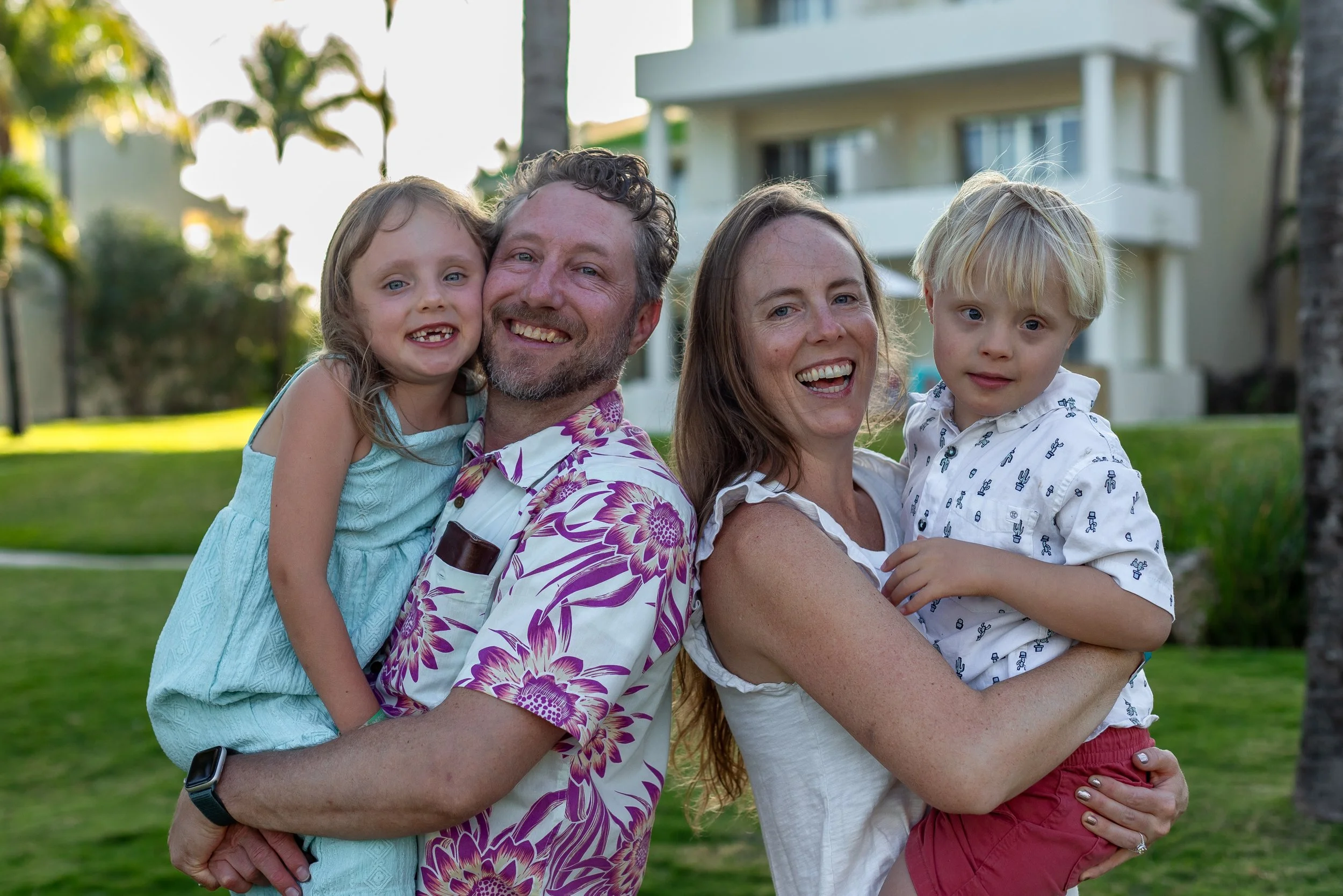A happy family of four outdoors with green grass, palm trees, and a white building in the background, smiling and posing for a photo.
