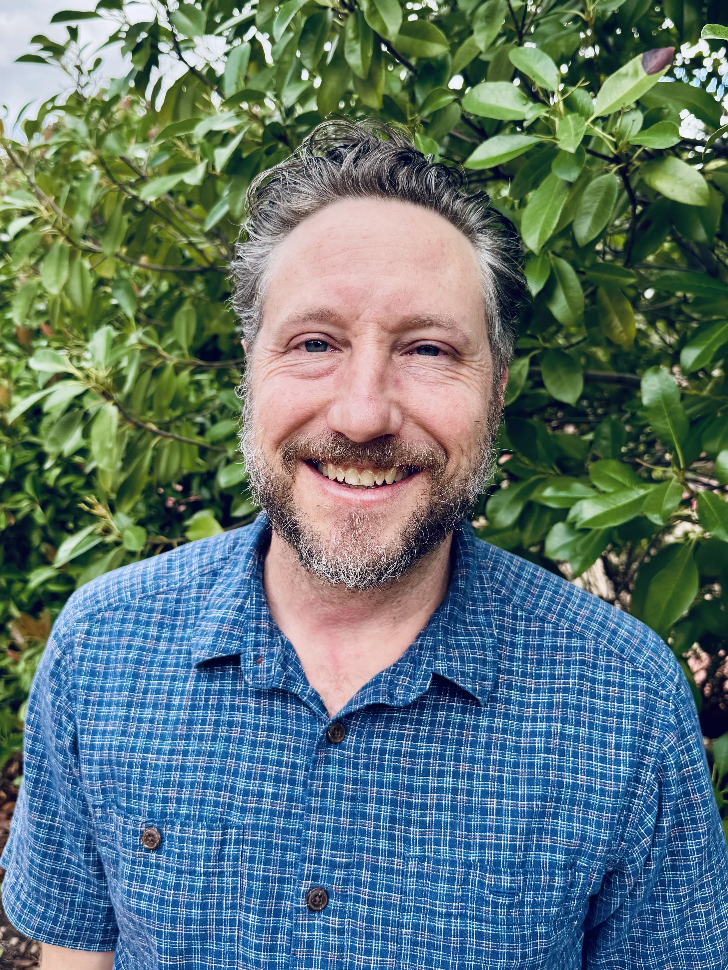 A smiling man with a beard and dark hair standing outdoors in front of green leafy bushes.