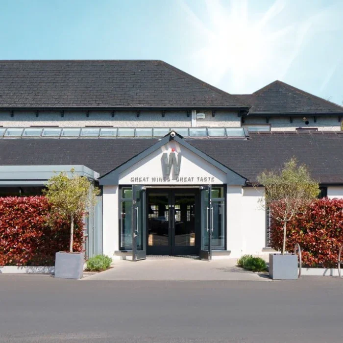 The entrance to a building with the sign 'Great Wines, Great Taste' above the door, surrounded by bushes and trees, with a clear sky above.