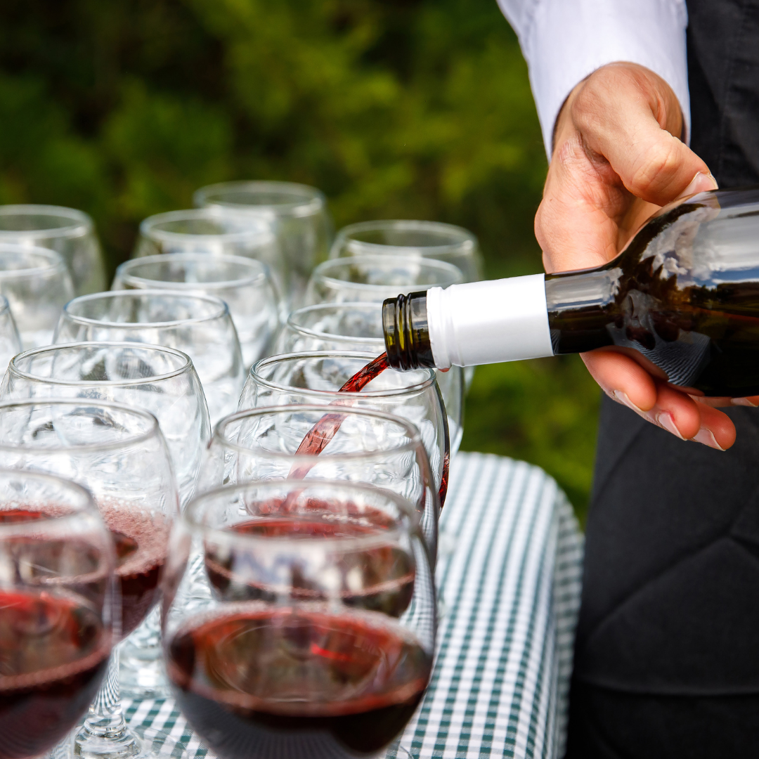 Man's hand pours red wine into a line of stemless wine glasses outside on a checked tablecloth