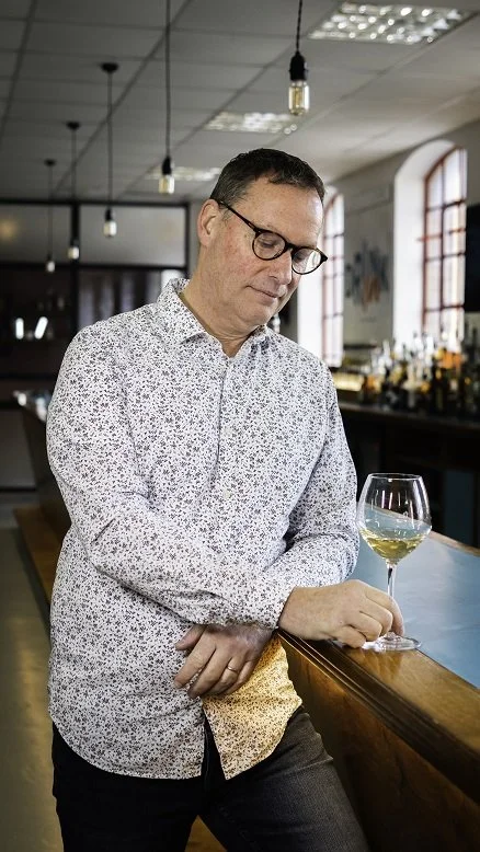 Man with glasses in a patterned shirt standing at a bar with a glass of white wine, in a dimly lit bar or restaurant.