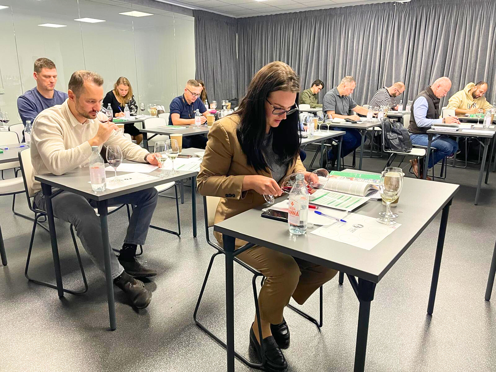 People seated at tables in a classroom, tasting wine, with glasses and bottles on the tables.