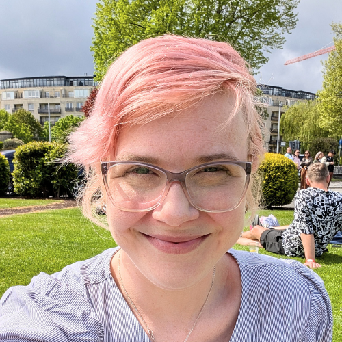 Headshot of woman with pink hair and glasses smiling at the camera