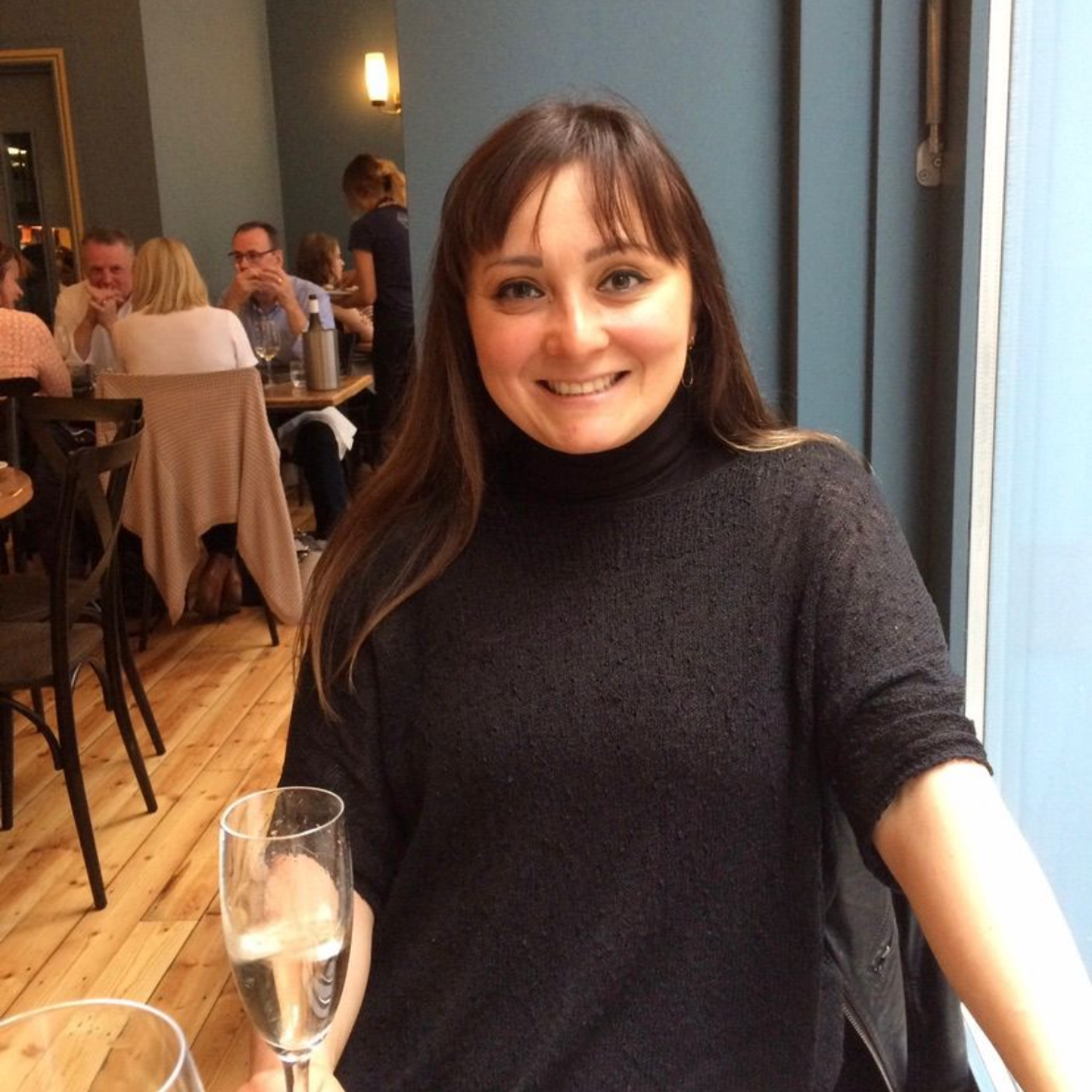 Woman with long dark hair and black top sitting at restaurant, holding a glass of sparkling wine