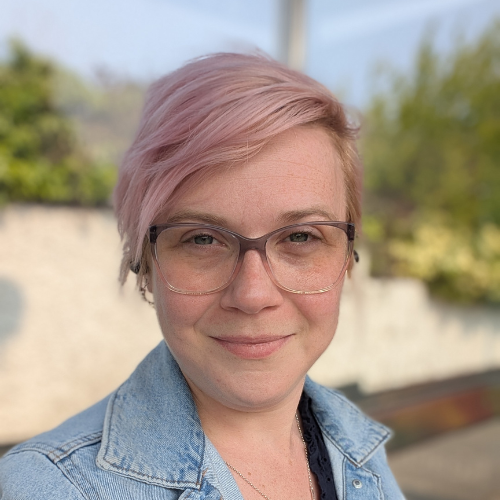 A woman with short pink hair, glasses, and a denim jacket smiling outdoors against a blurred background of trees and buildings.