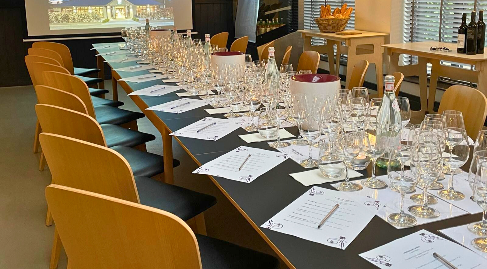 A long dining table set with wine glasses, water glasses, silverware, and printed menus. There are empty chairs around the table, with a black tablecloth. In the background, there are wine bottles, baskets of bread, and a projection screen.