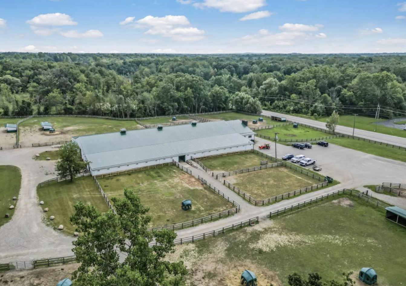Aerial view of a rural farm with a large barn, fenced animal pens, parking lot, and surrounding green trees under a partly cloudy sky.
