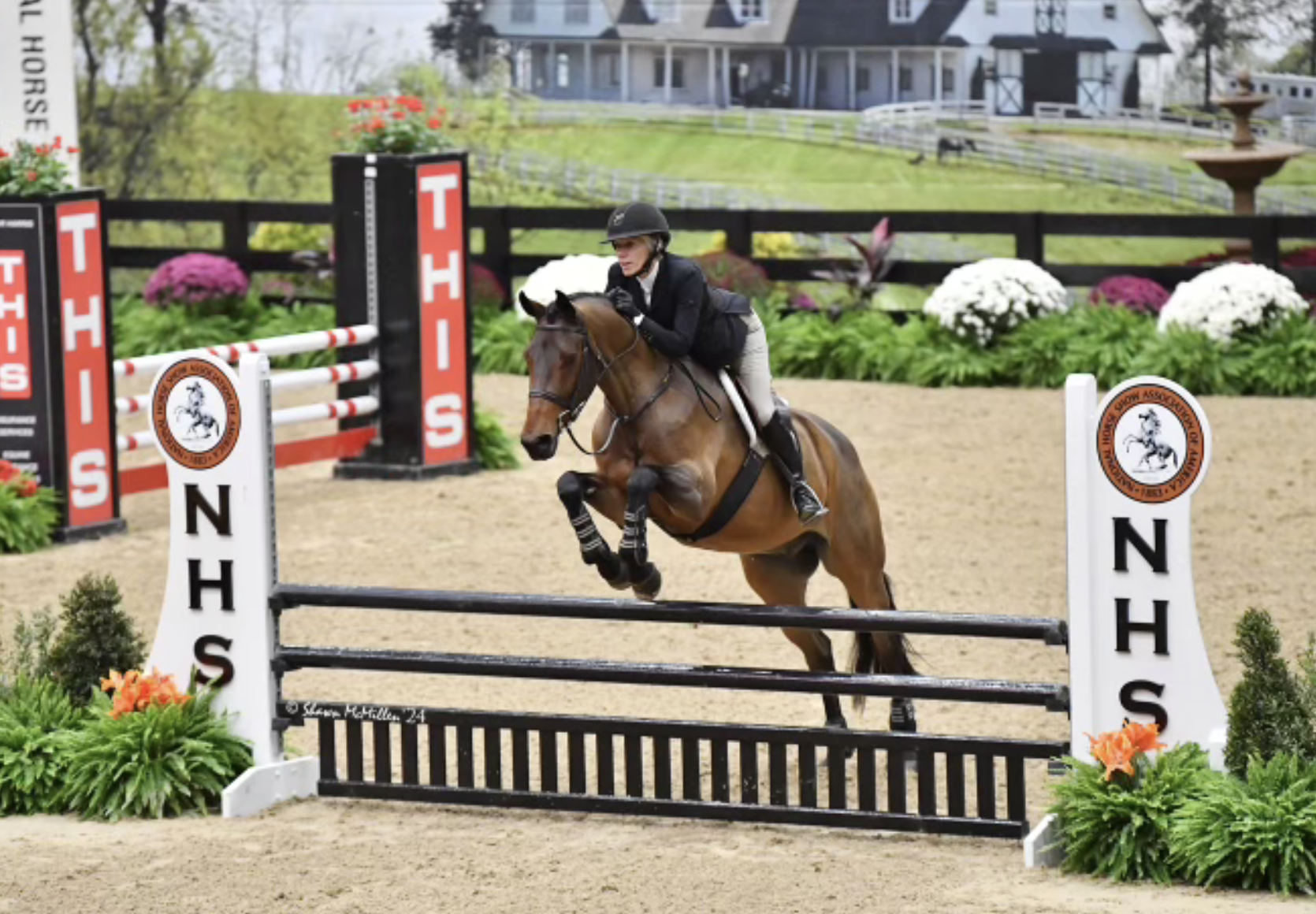 A female equestrian riding a brown horse over a jump in a show jumping competition. The jump is labeled with 'NHS' and is decorated with flowers.