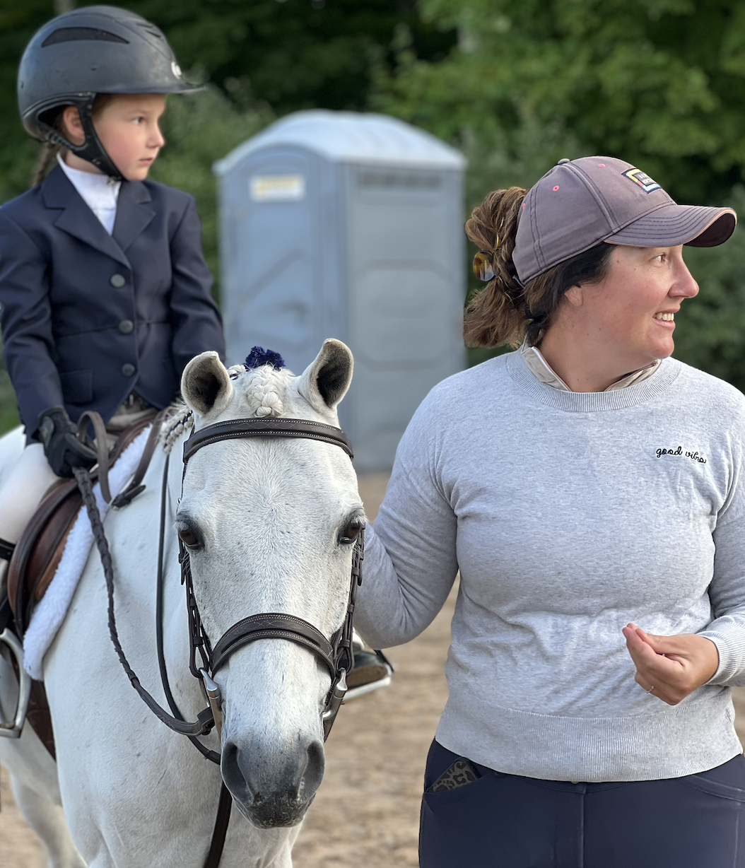 A woman smiling and holding the reins of a white horse with a young girl riding it, wearing a black riding helmet and a navy blazer, outdoors with a portable toilet and trees in the background.