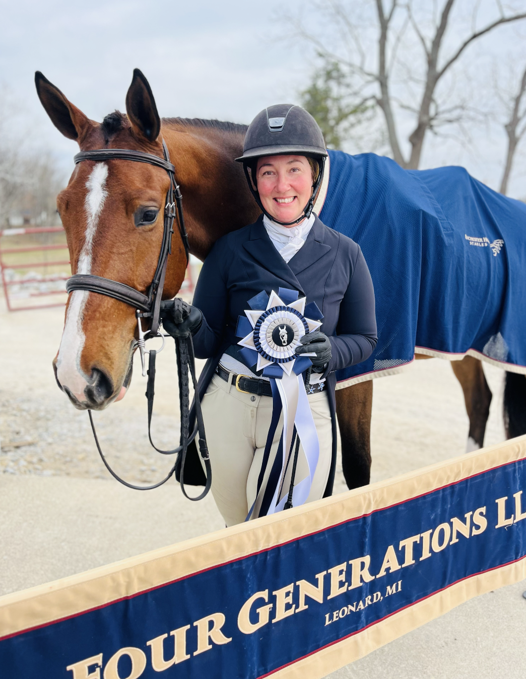 A woman in equestrian attire smiling while holding a blue and white ribbon next to a brown horse wearing a blue blanket. They are outdoors near a sign that says "FOUR GENERATIONS LLC LEONARD, MI."