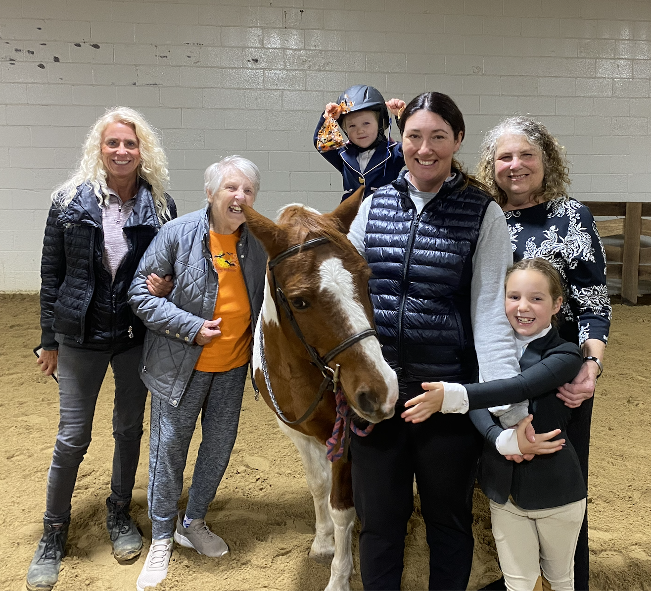 Group of six women and two children posing with a brown and white horse in an indoor riding arena, some smiling and hugging, one child wearing a helmet, the women wearing casual jackets and sweaters.