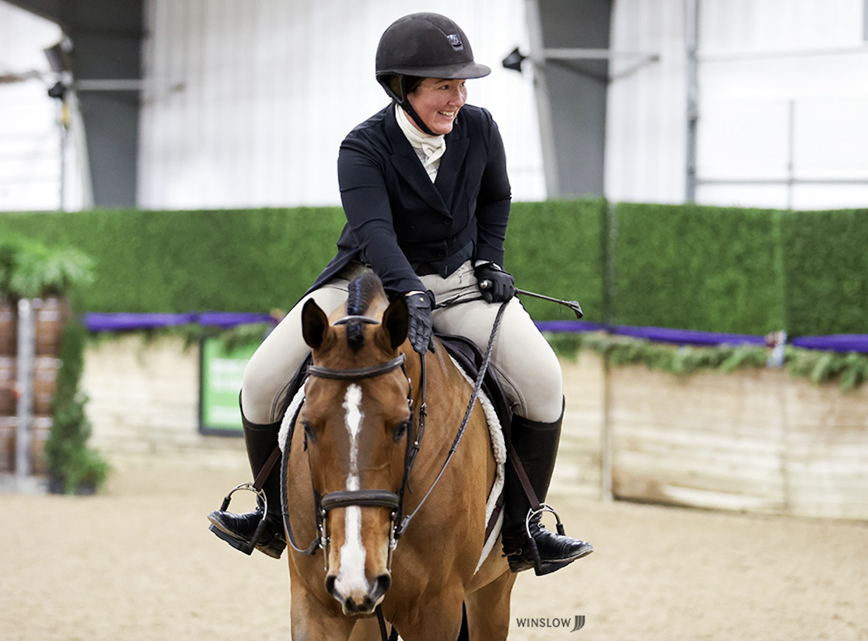 A woman wearing a black riding helmet, black riding coat, and beige jodhpurs riding a chestnut horse with a white blaze in an indoor riding arena, smiling.