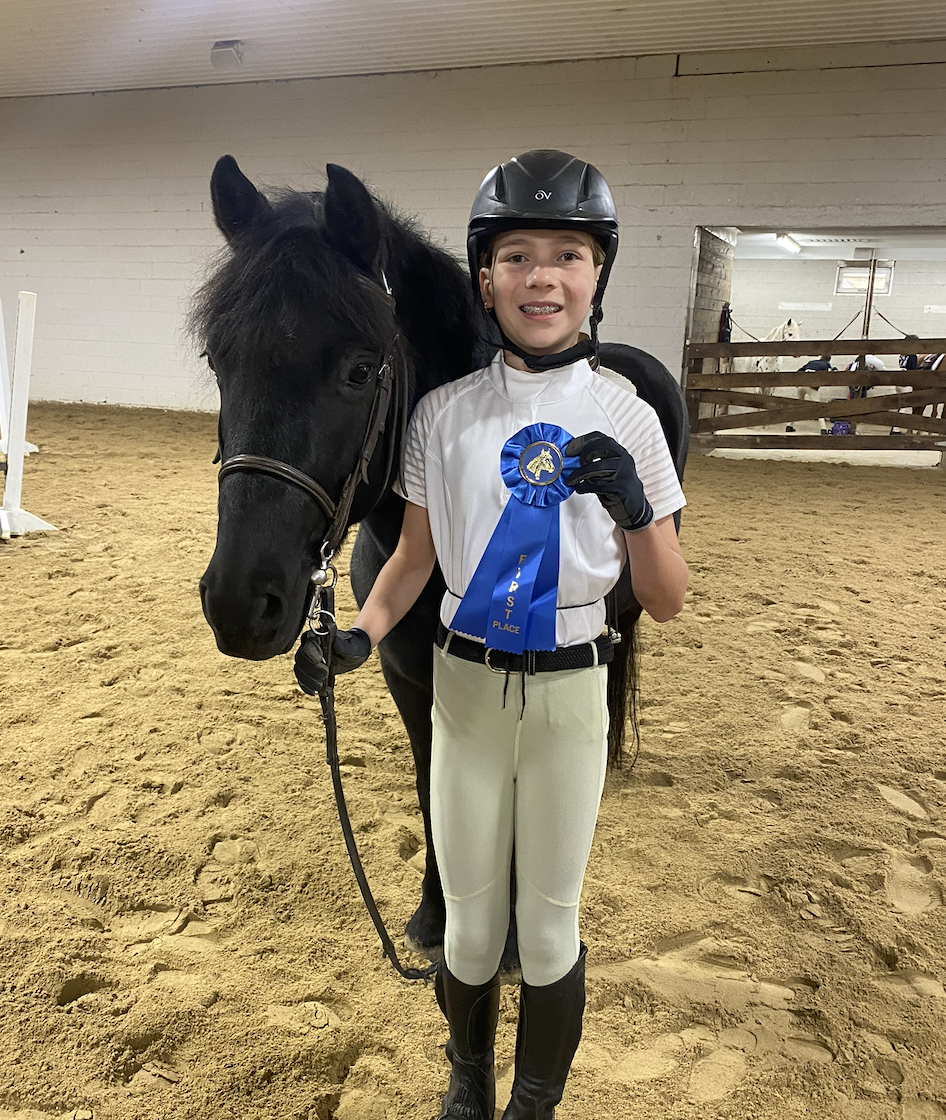 A young girl in equestrian attire holding a blue ribbon, standing next to a black horse in an indoor riding arena.