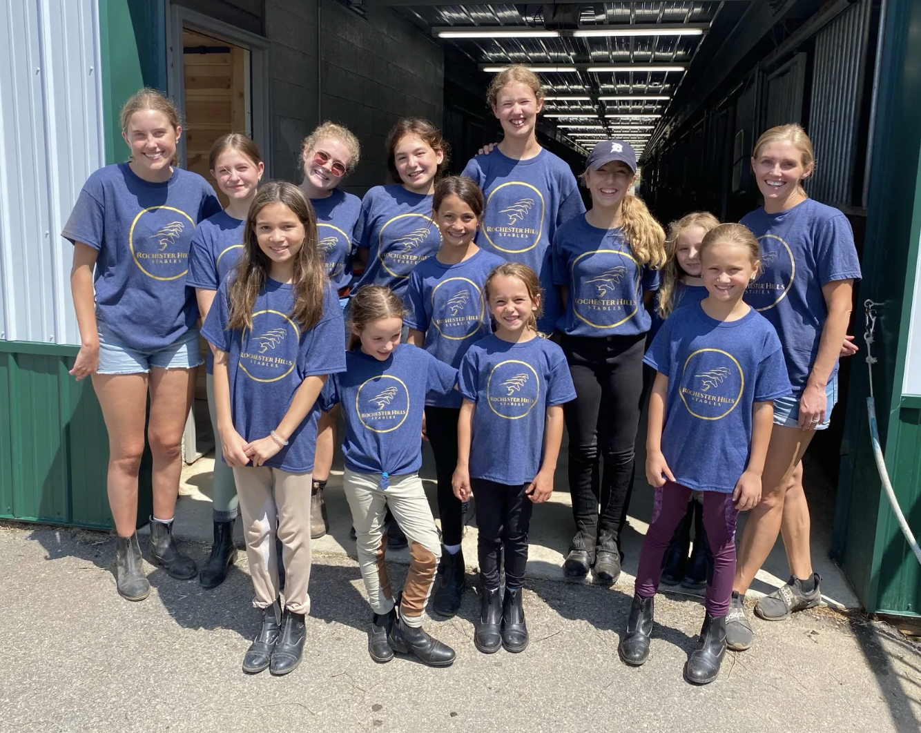 Group of girls and women wearing blue Rochester Hills Stables t-shirts standing together outdoors in front of stables, smiling at the camera.