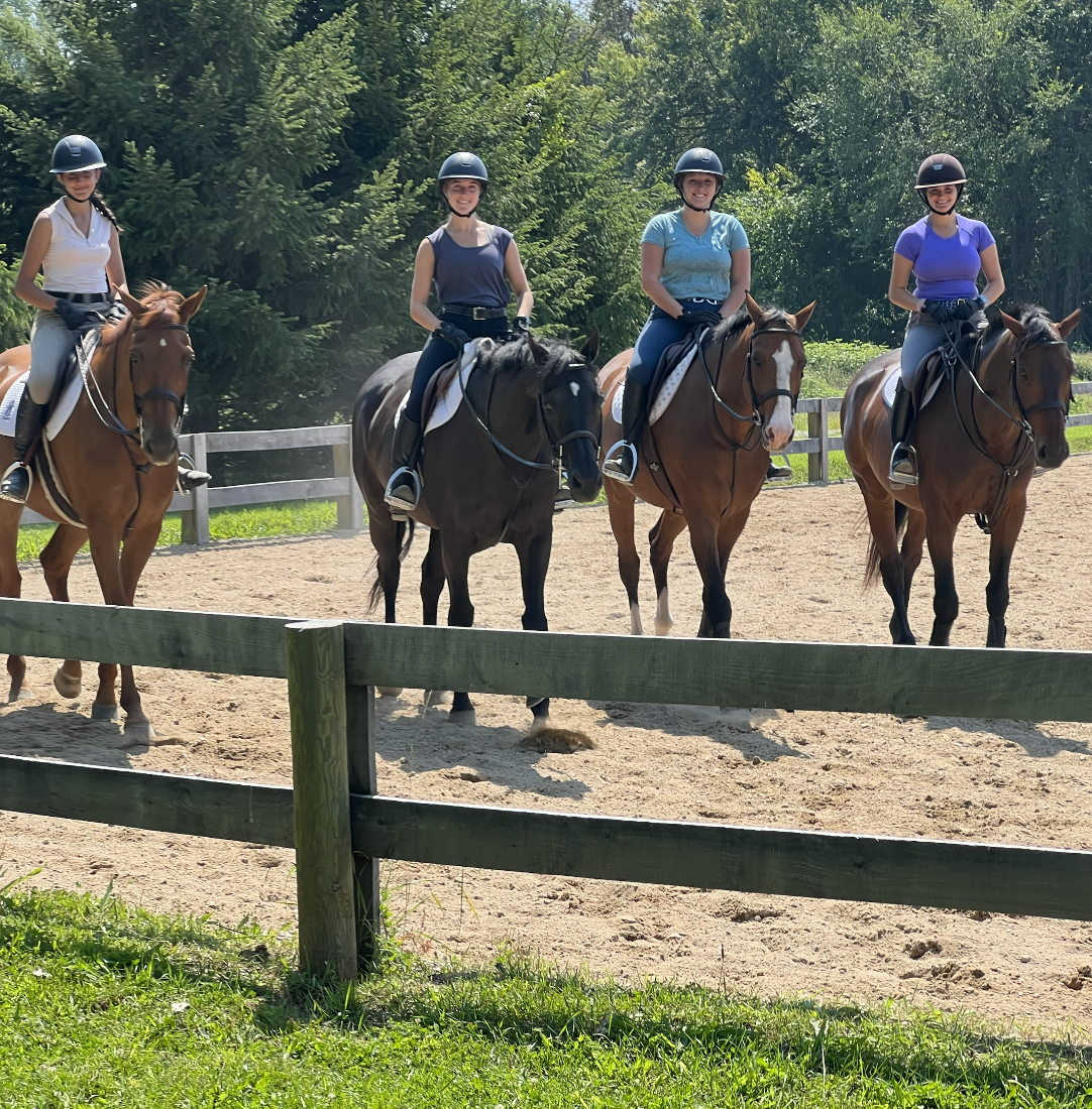 Four women riding horses in an outdoor riding arena, all wearing helmets and riding casual clothing, with a wooden fence and green trees in the background.