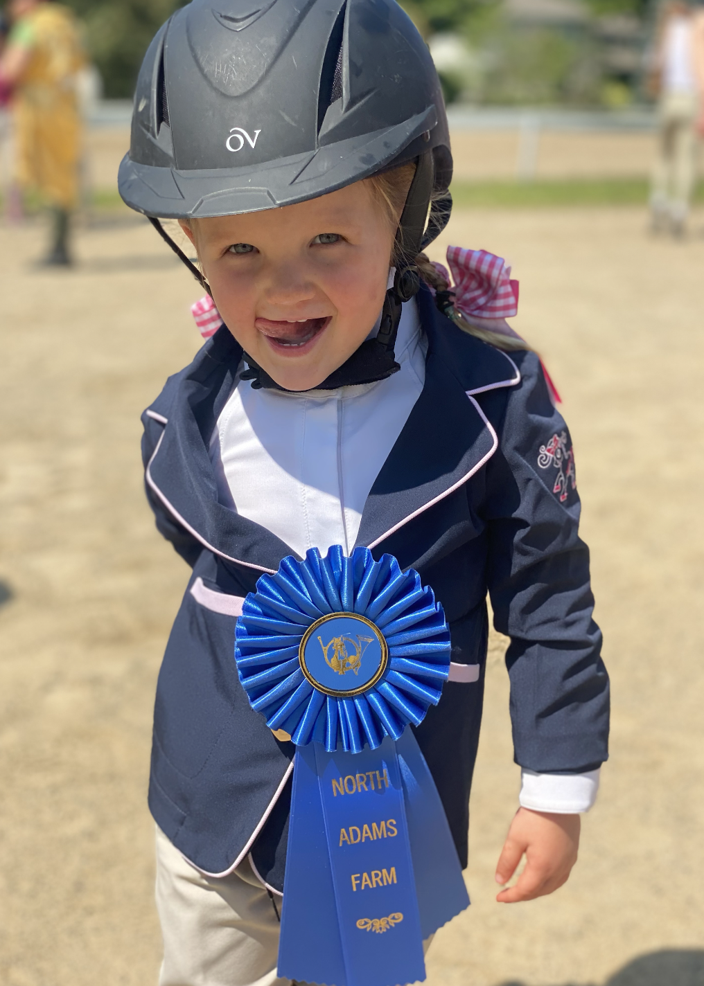 A young girl wearing a black riding helmet, a navy riding jacket with pink piping, and a white shirt. She has a blue ribbon with gold text that says 'North Adams Farm' pinned to her jacket. The girl is smiling and leaning forward, with a colorful outdoor background.