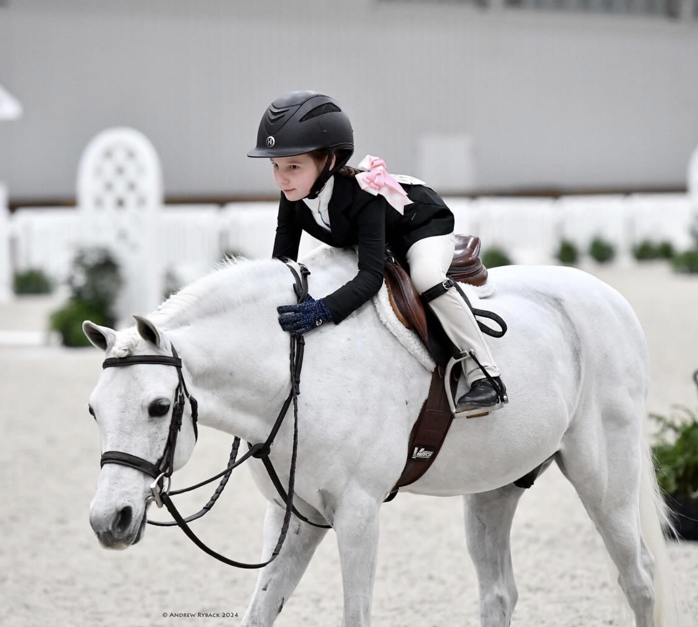 Young girl riding a white horse indoors, wearing a black helmet and riding gear.