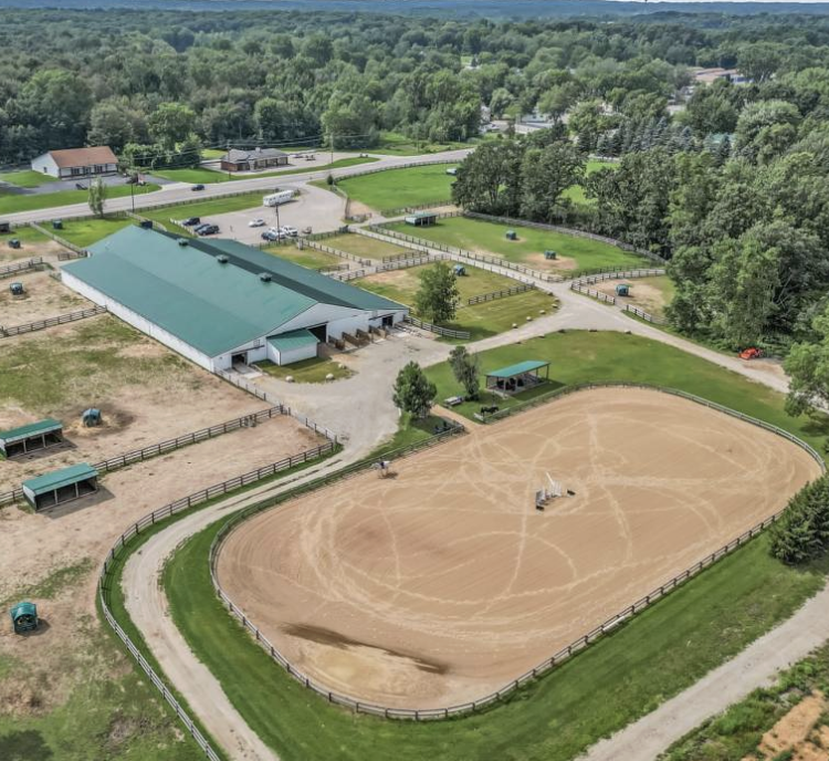 Aerial view of an outdoor equestrian facility with a large riding arena, stables, and surrounding trees.