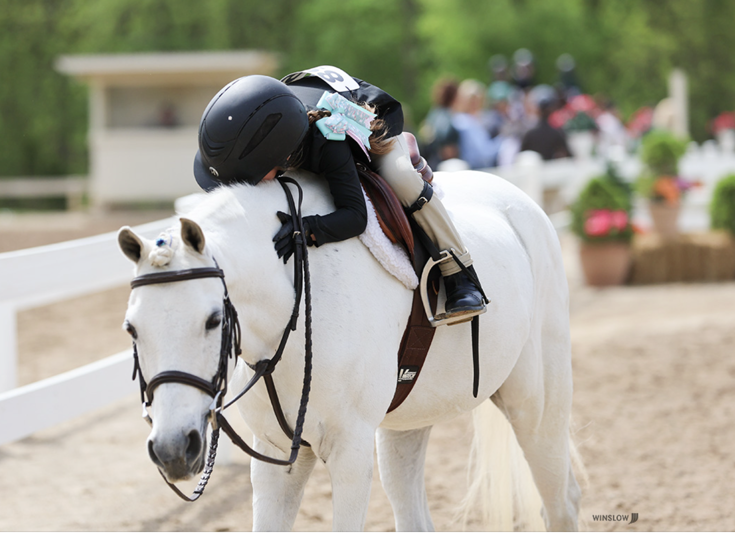 Young girl leaning forward, hugging a white horse's neck, wearing a black riding helmet and black riding outfit, at an outdoor equestrian event with a sandy riding arena and blurred spectators in the background.