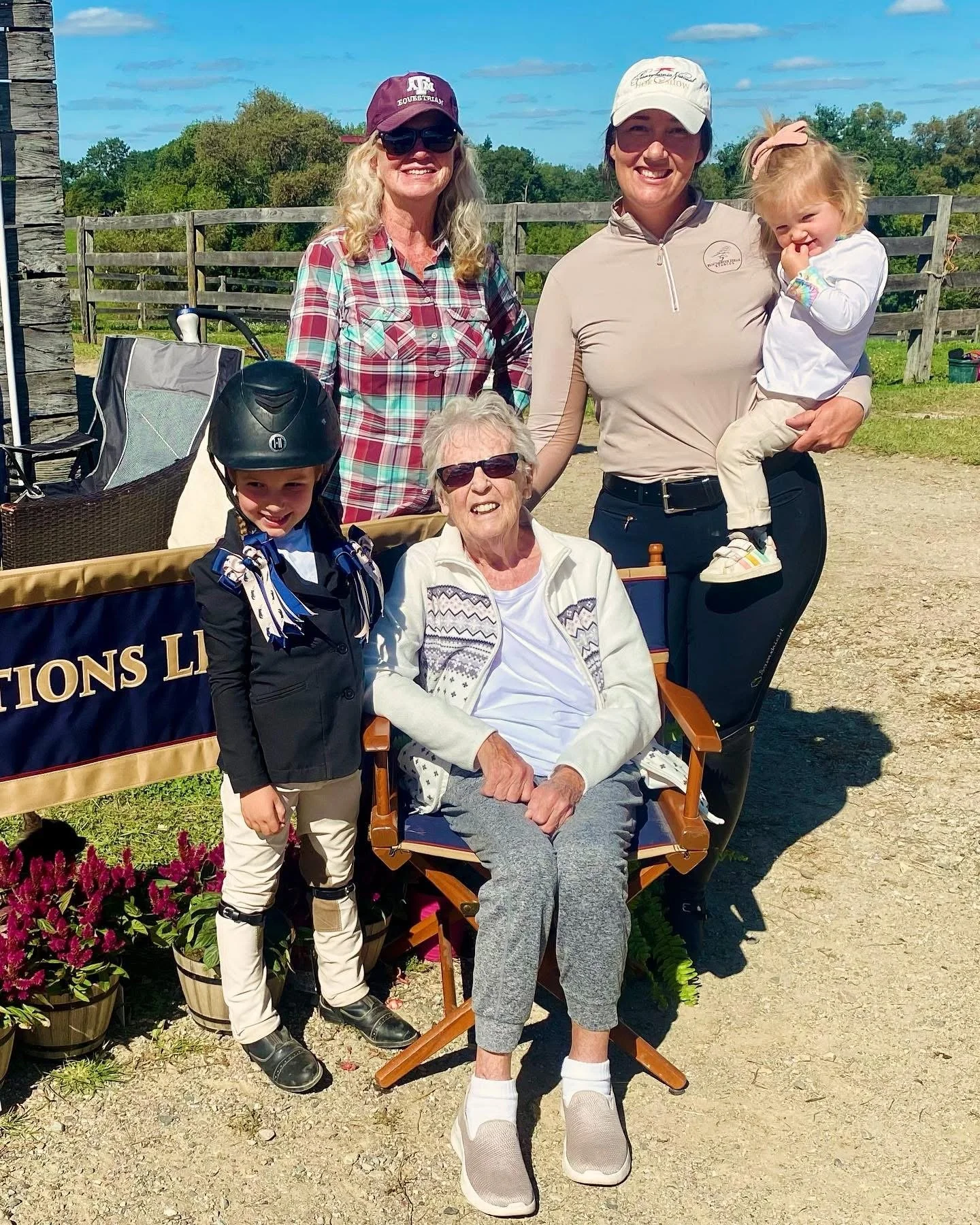 Group of five women and two children outdoors on a sunny day, with a wooden fence and greenery in the background. One woman is sitting on a folding chair, others are standing, one girl is wearing a helmet, and the youngest girl is being held by a woman, all smiling.