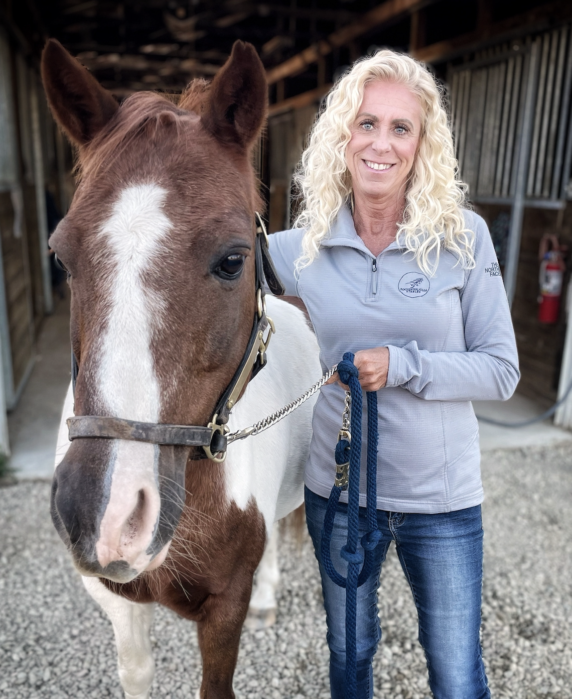 A woman with curly blonde hair smiling next to a brown and white horse inside a stable.
