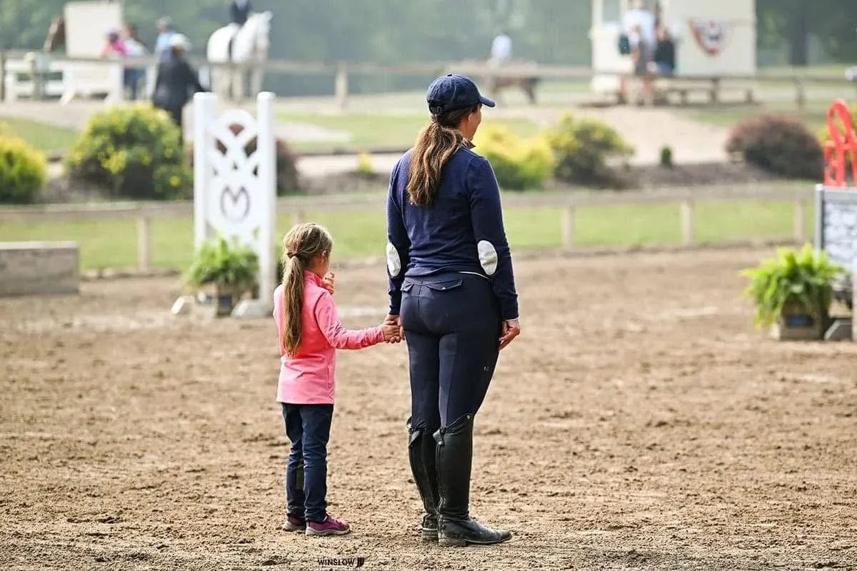 A young girl holding hands with an adult woman, likely her mother, at an outdoor equestrian riding arena. The girl is wearing a pink jacket and jeans, while the woman is dressed in equestrian attire, including riding boots, breeches, a navy jacket, and a cap. In the background, there are some people, greenery, and equestrian jumps.