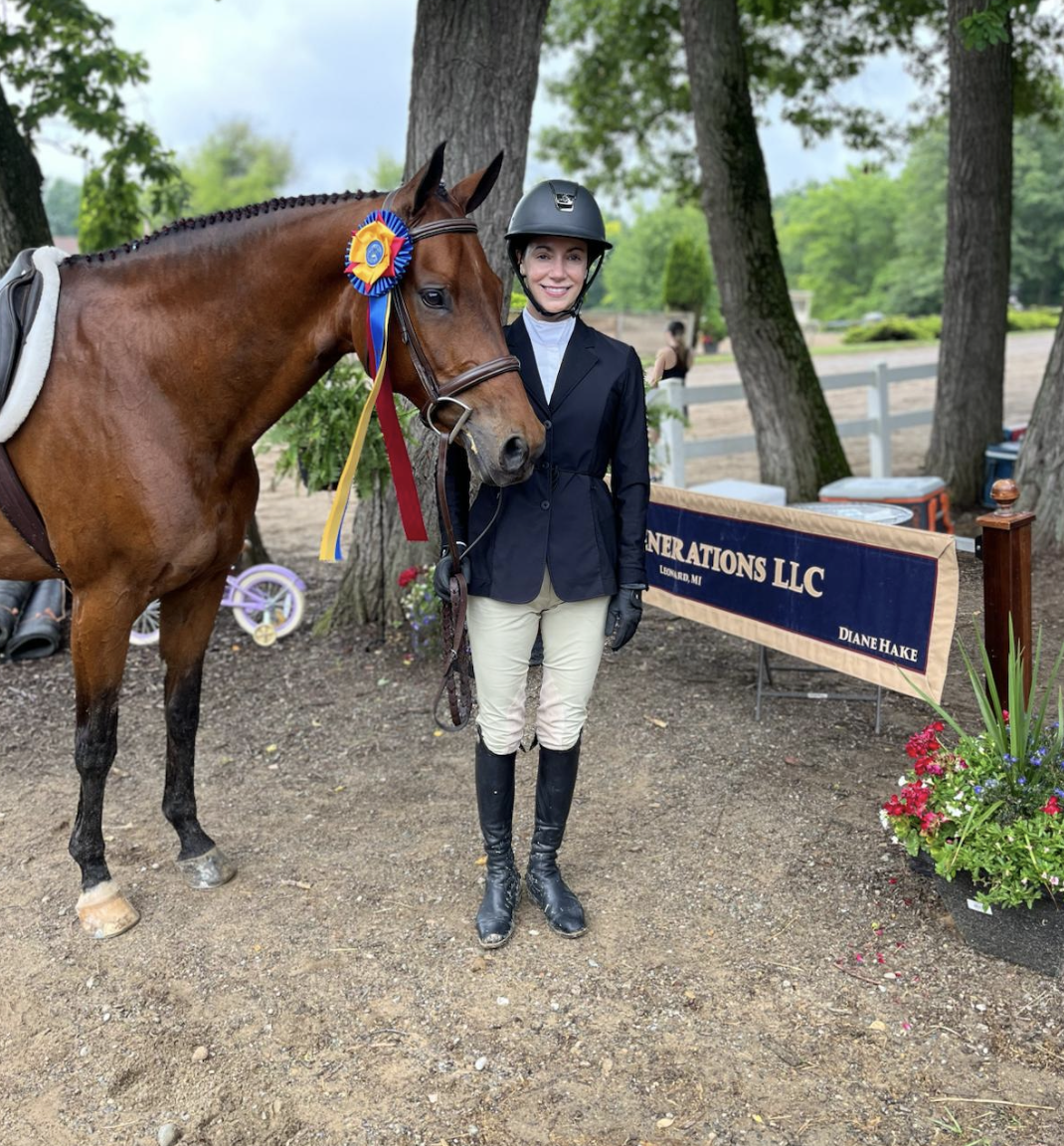 A woman in equestrian attire, including a helmet, standing next to a brown horse decorated with colorful ribbons, outdoors near trees and a white fence, with a ribbon and a banner in the background.