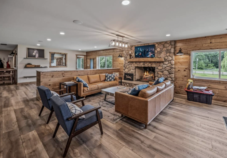 A cozy living room with wooden walls and flooring, featuring a stone fireplace with a TV above. There are three sofas and two armchairs arranged around a coffee table, with decorative pillows. Large windows show greenery outside, and a ceiling light fixture is visible.