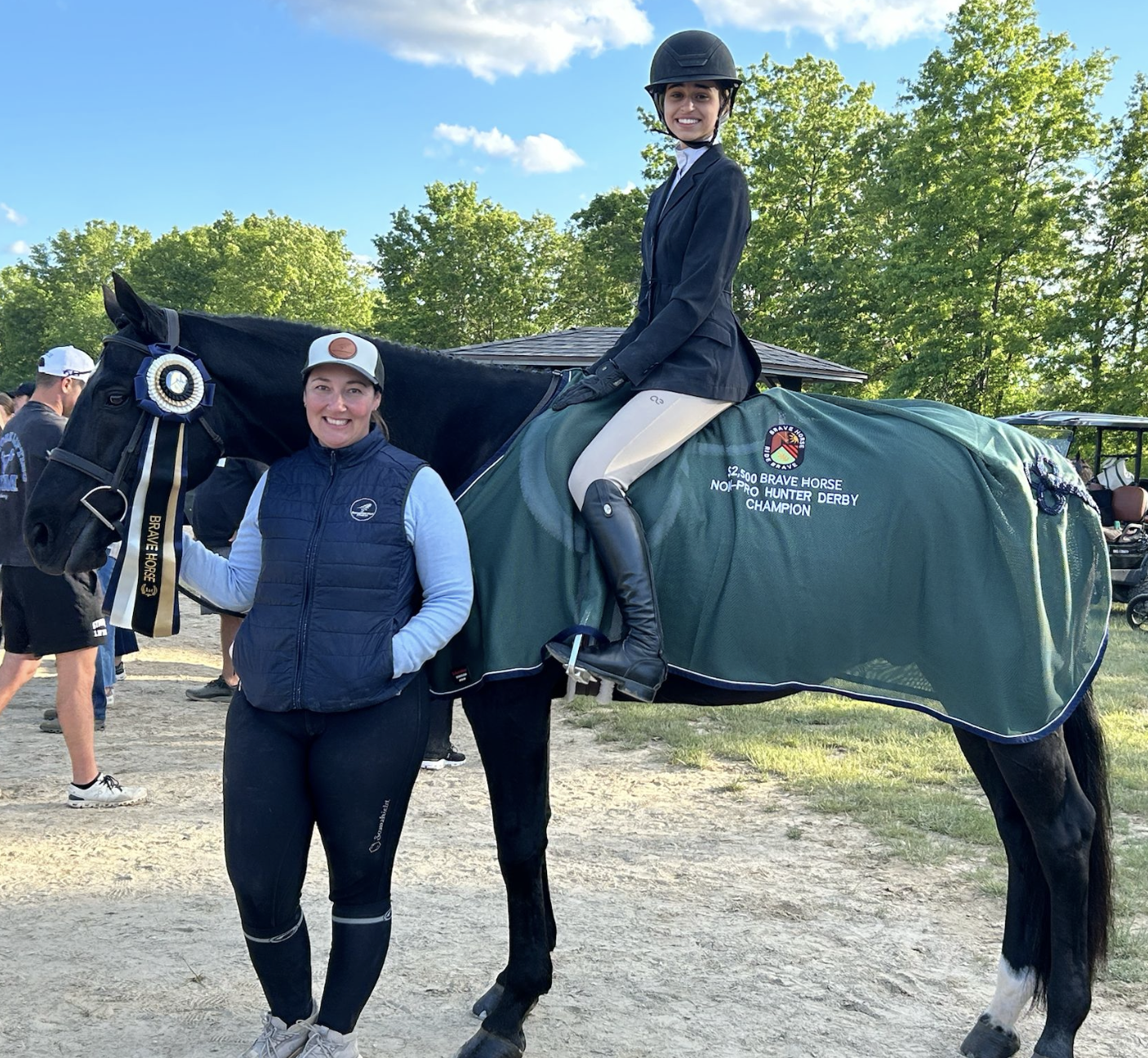 A woman standing next to a black horse with a ribbon and a rider on the horse, who is wearing riding attire and a helmet, at a horse racing event.