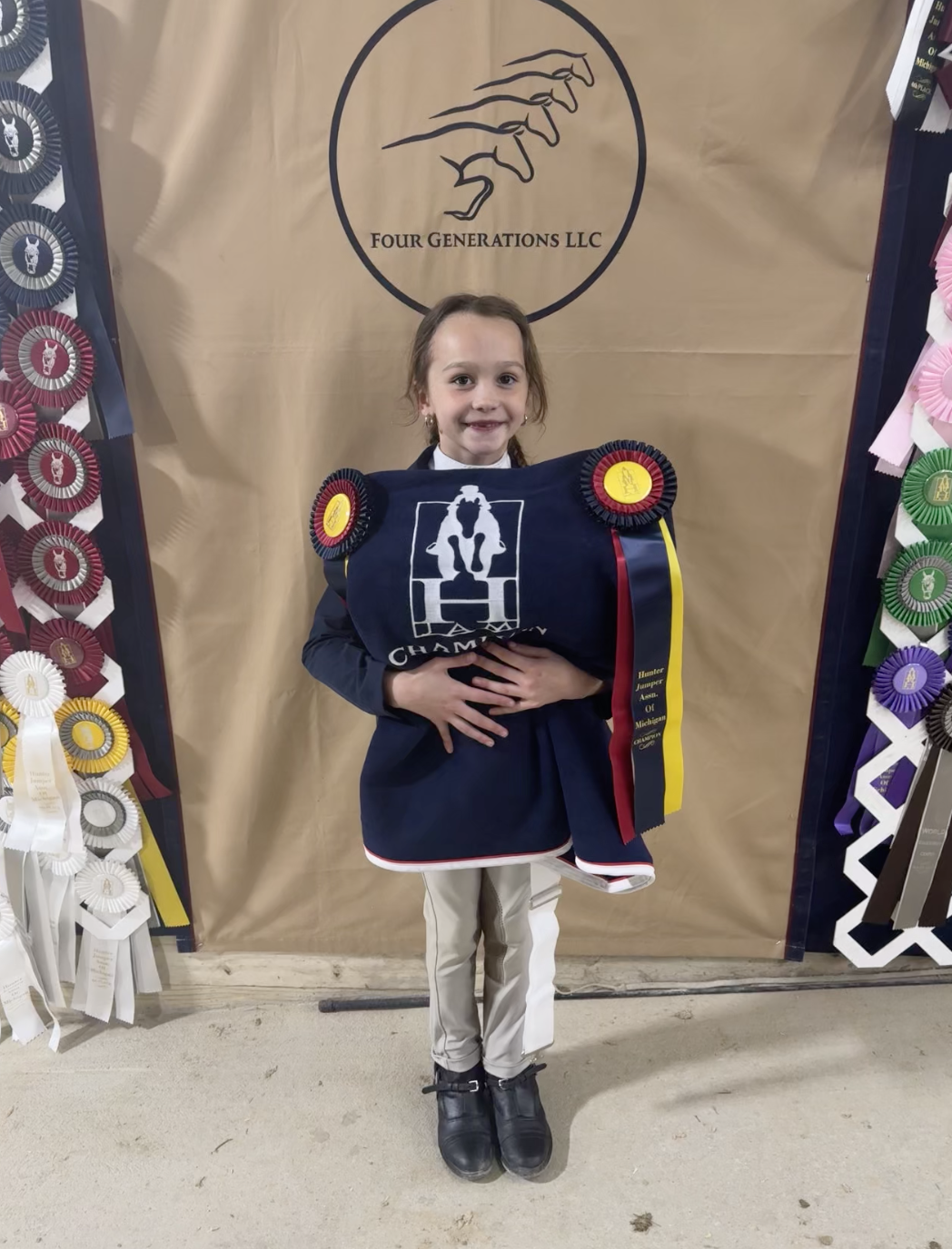 A young girl holding a championship blanket and ribbons, standing in front of a beige backdrop with a logo of four stylized hands and the text 'Four Generations LLC,' surrounded by various award ribbons.