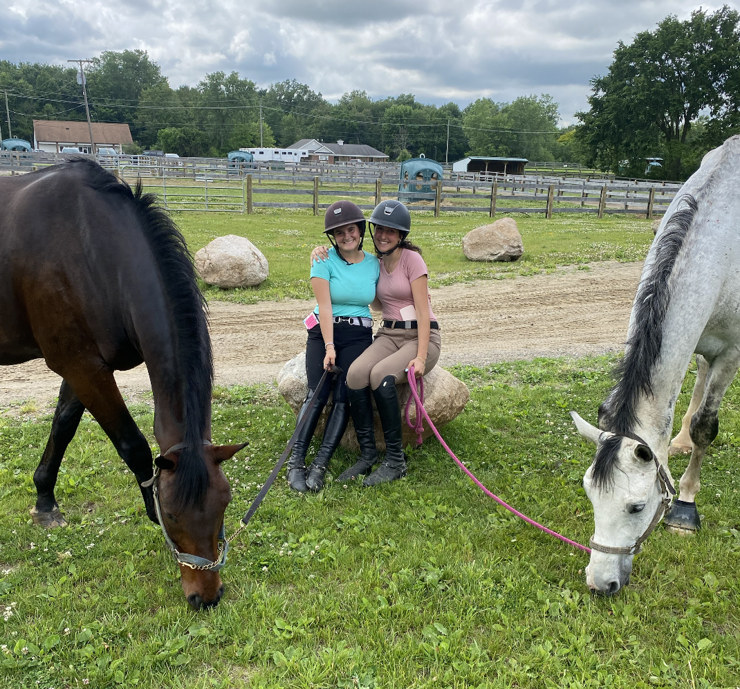 Two women in riding helmets sitting on a large rock with their horses grazing nearby in a rural field with fencing, trees, and a cloudy sky in the background.