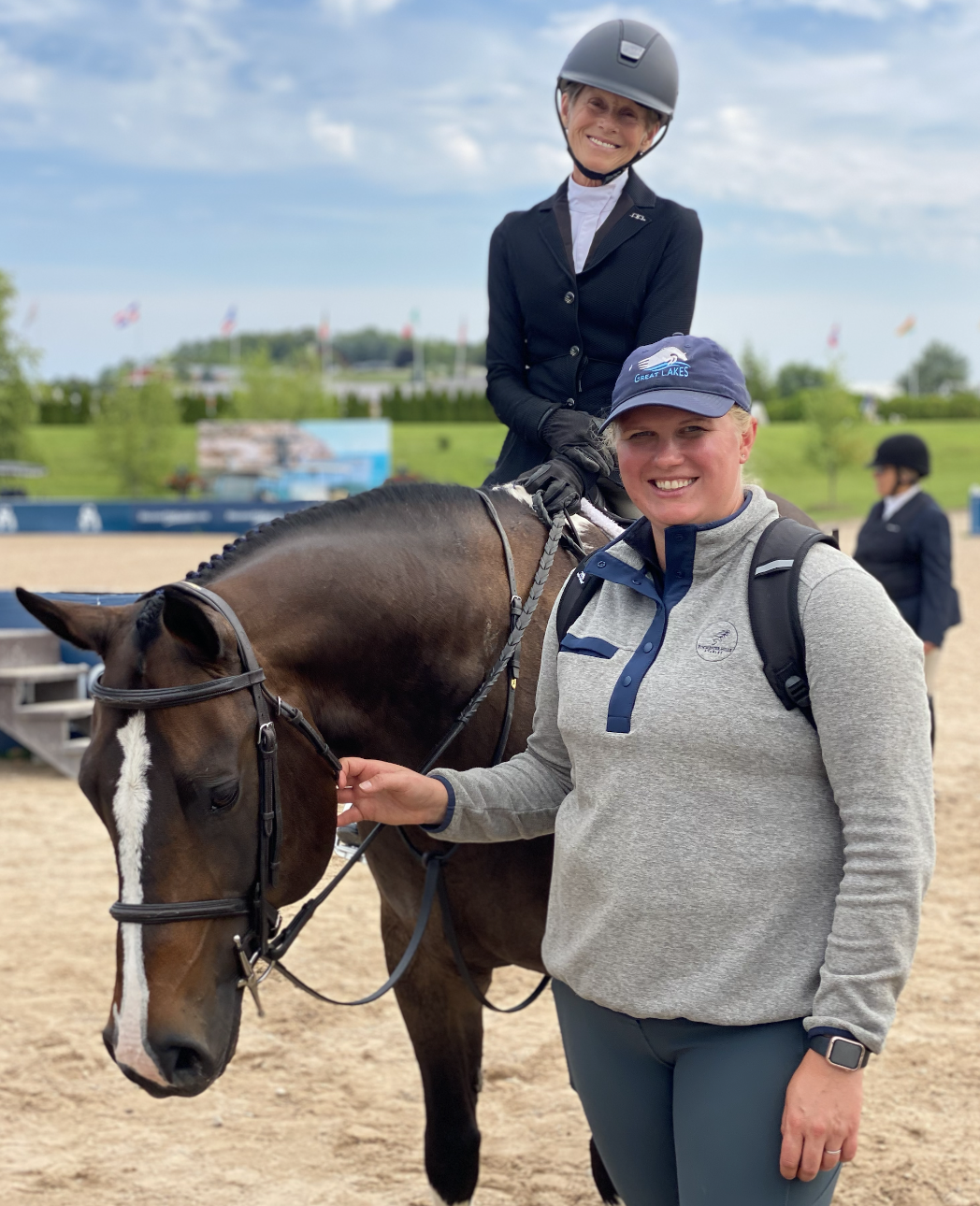 Two women and a horse at an outdoor equestrian event, with one woman sitting on the horse and the other standing beside it, holding the reins.