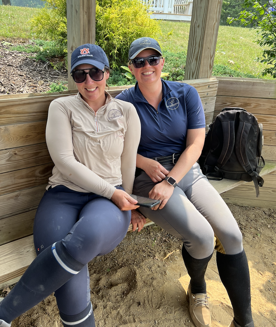 Two women dressed in equestrian riding attire, sitting on a wooden bench outdoors, smiling at the camera. One woman is wearing a beige quarter-zip top, navy riding pants, and a navy cap with an Auburn logo, holding a phone. The other is dressed in a navy riding shirt, light gray riding pants, black riding boots, a cap, and sunglasses. A black backpack is on the bench beside them, with greenery and a small garden in the background.