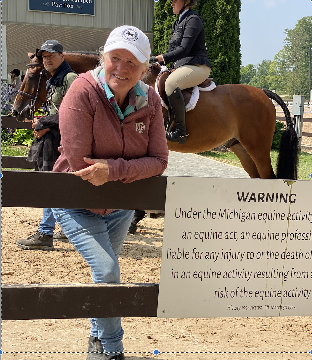 A woman in a red jacket leaning on a fence at a horse riding event, with a smiling face, wearing a white cap with a logo. Behind her, a young rider in riding attire is seated on a brown horse, with another rider and a man with a horse in the background during daytime. There is a warning sign about equine activity liability.