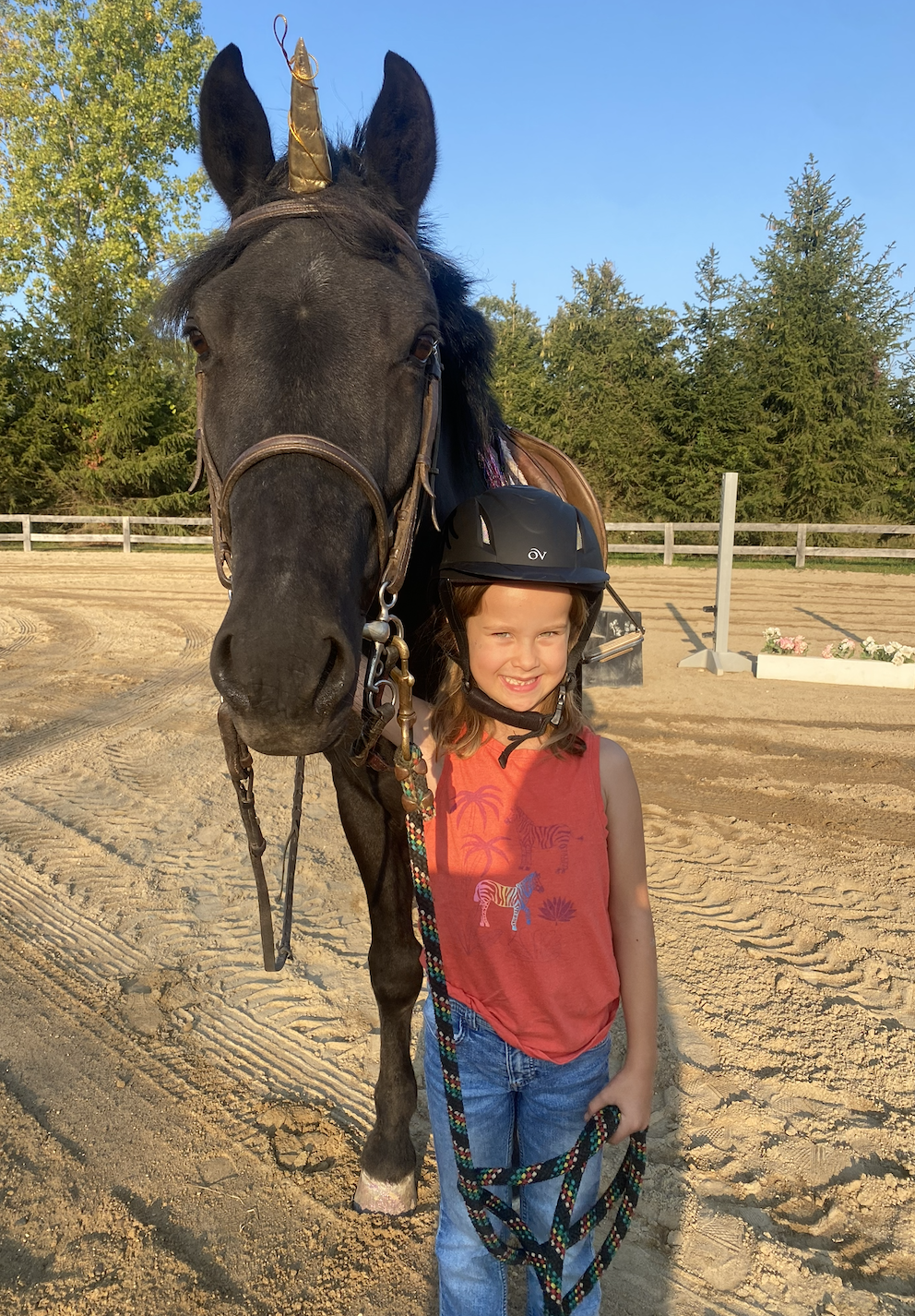 A smiling young girl wearing a black riding helmet and a sleeveless orange shirt with zebras and palm trees stands next to a large black horse with a golden horn, which appears to be a unicorn. They are outdoors on a dirt farm or riding arena, with trees and a fence in the background, during late afternoon or early evening sunlight.