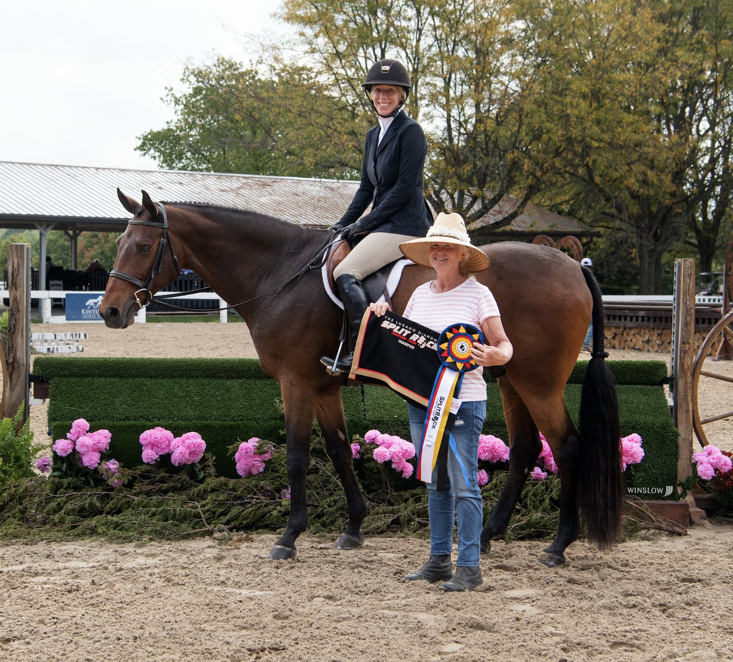 A woman in a riding helmet and equestrian outfit riding a horse, with another woman standing beside them holding a ribbon and rosette, on a decorated equestrian show ring with pink flowers and a dirt arena.