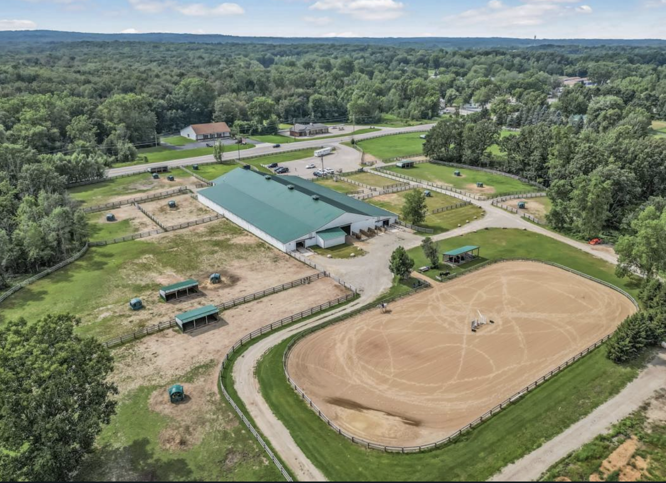Aerial view of a horse riding facility with an indoor riding arena, paddocks, and a large outdoor riding arena surrounded by fences, with trees and greenery in the background.