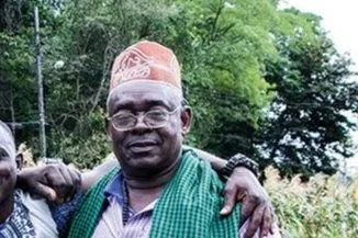 A man wearing glasses and a traditional pink hat with a green scarf outdoors with trees in the background.