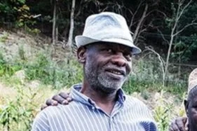 A man outdoors wearing a light-colored fedora hat and striped shirt, smiling with a background of greenery and trees.