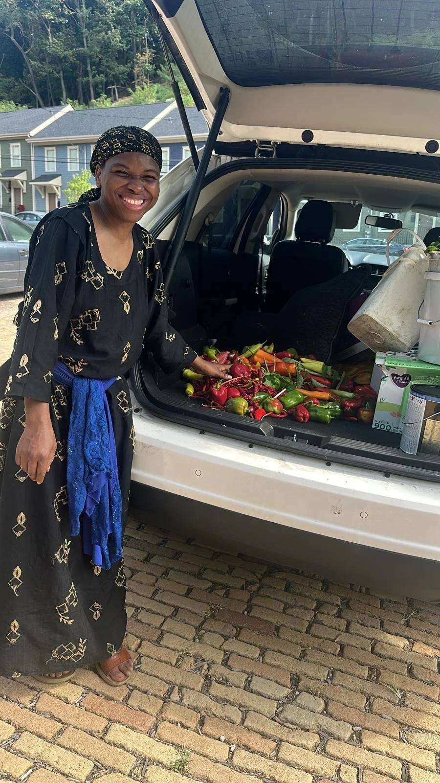A woman smiling and wearing a headscarf and a black dress with gold patterns, standing next to an open trunk filled with freshly harvested peppers and carrots. The car is parked on a brick driveway with trees and houses in the background.