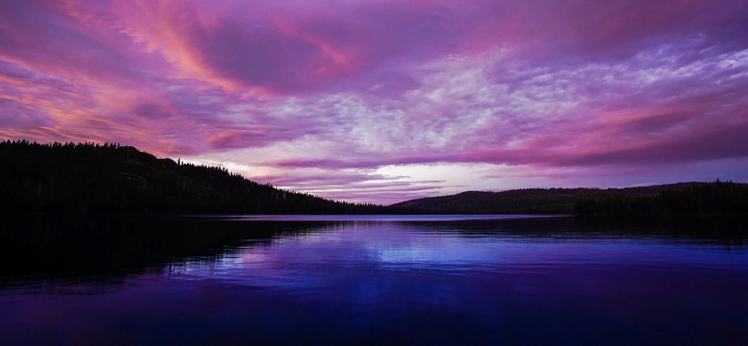 A landscape scene of a lake at sunrise, with a sky filled with pink and purple clouds, reflecting on the calm water, and forested hills in the background.