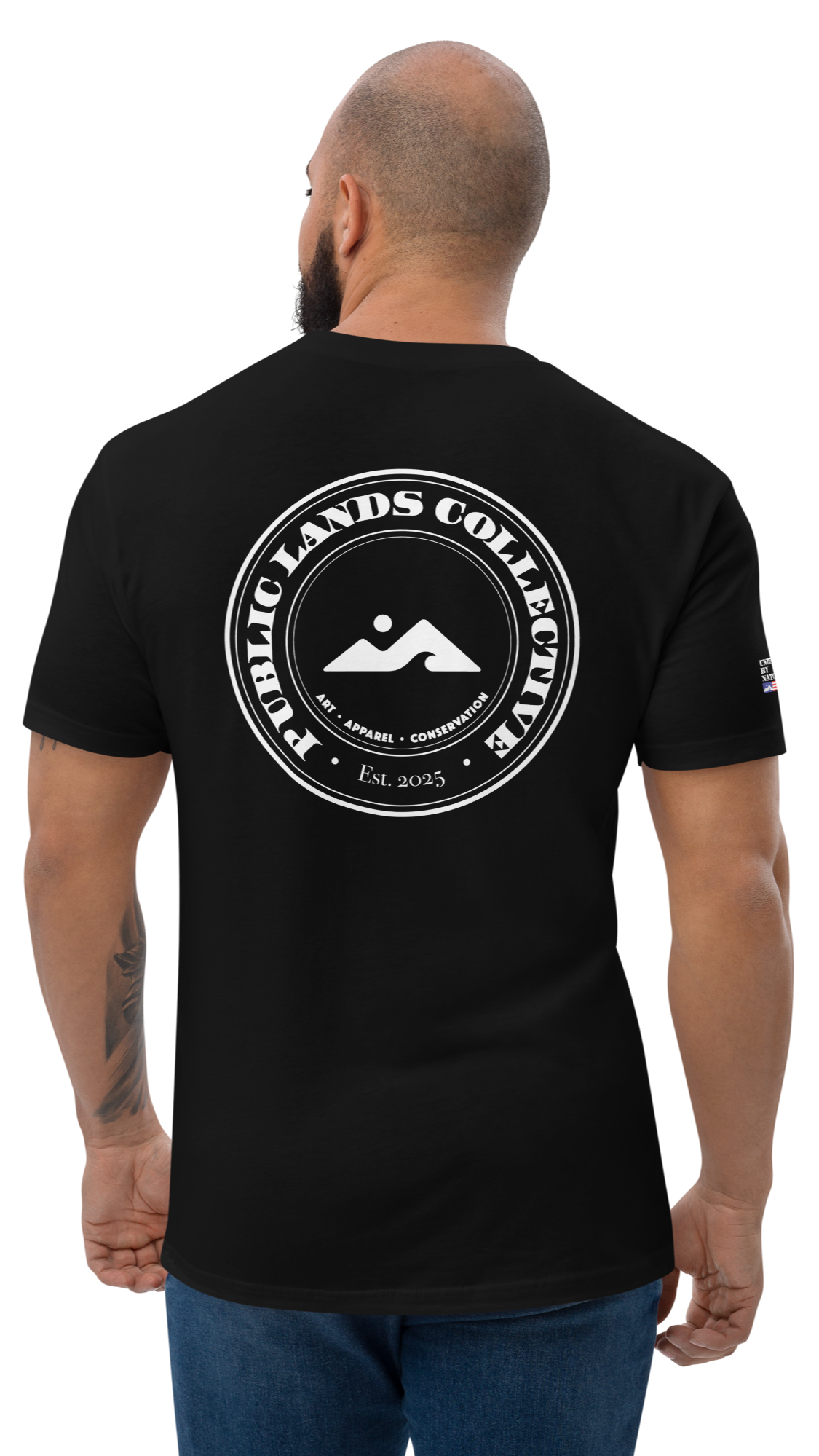 Back view of a man wearing a black t-shirt with a circular logo that reads "Public Lands Collective" and features mountain and sun graphics, with additional text "Art, Apparel, Conservation" and "Est. 2025."