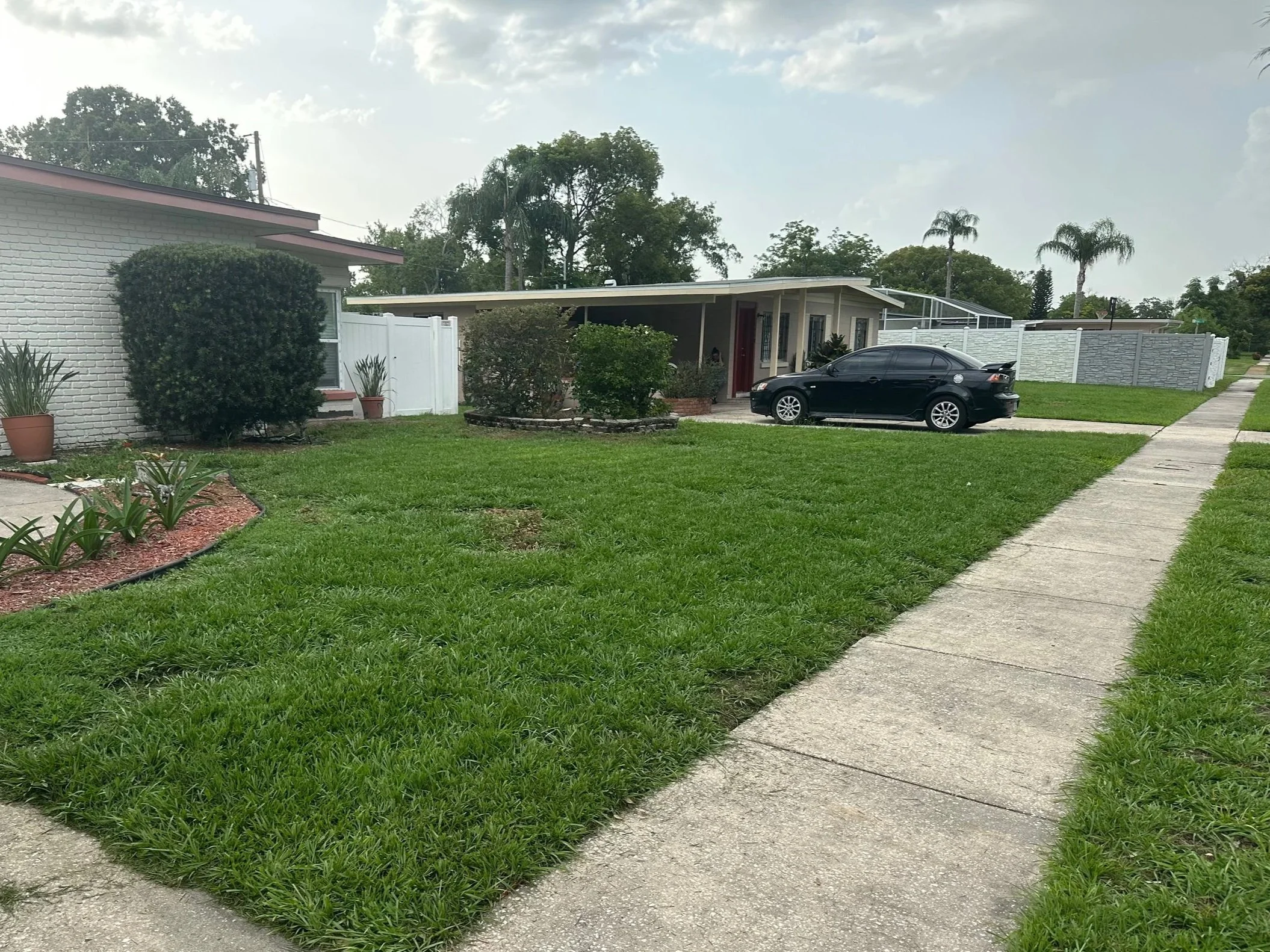 Front yard with a sidewalk, green grass, potted plants, and a house with a car parked in the driveway.