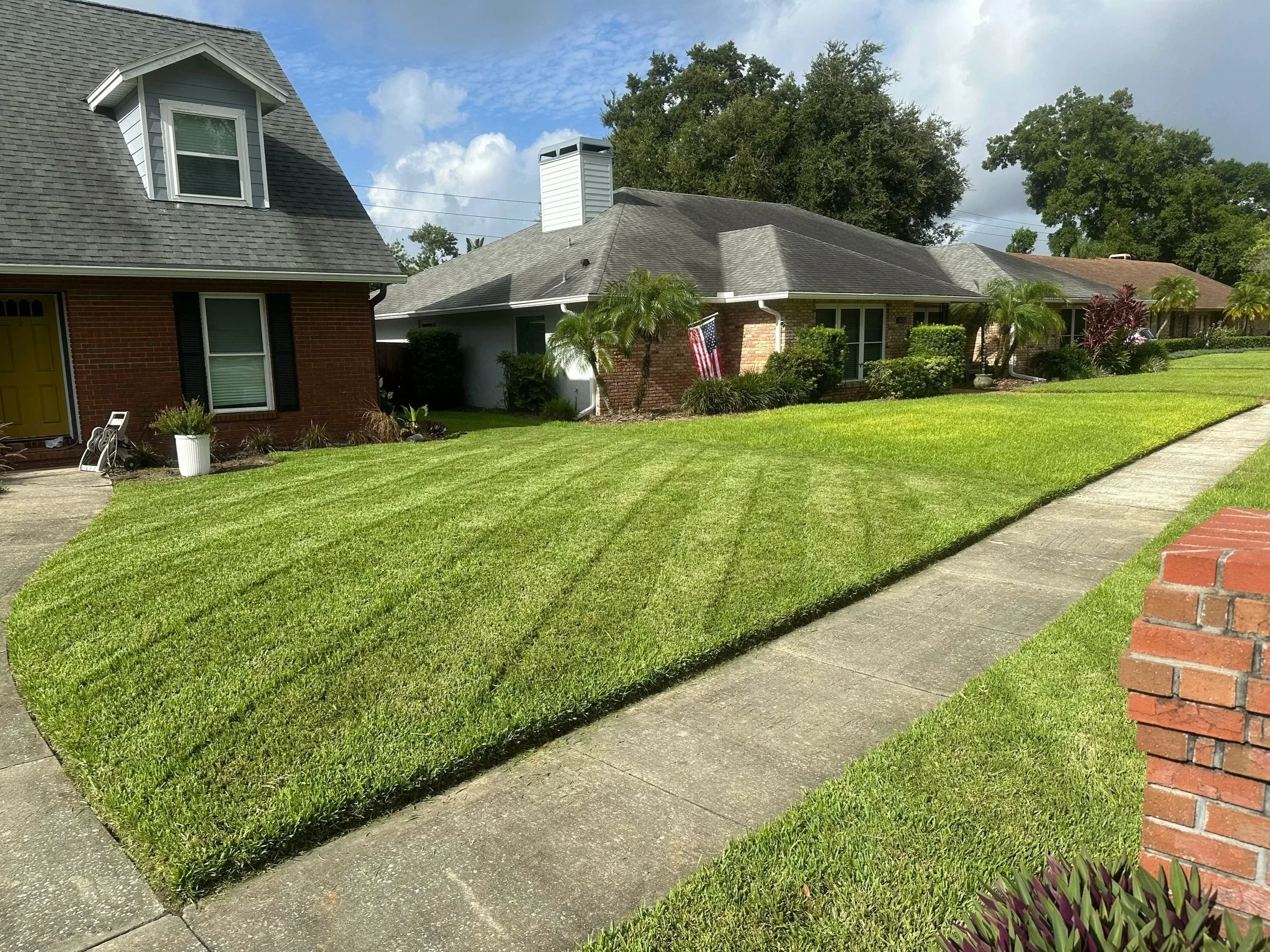 A well-maintained front lawn with striped grass, a sidewalk, and houses with brick and siding exteriors, trees, and bushes under a partly cloudy sky.