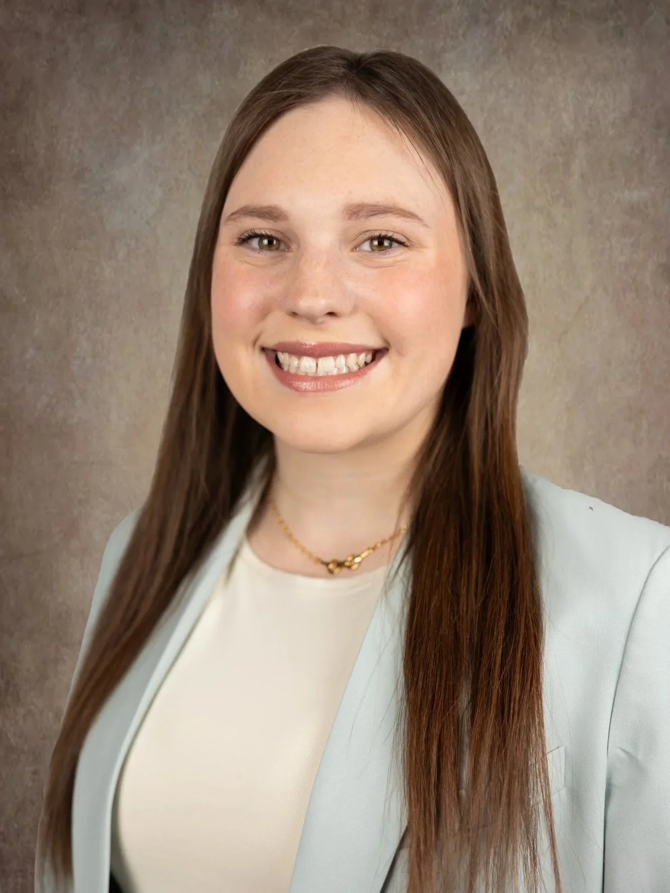 A young woman with long brown hair smiling, wearing a white top, a light-colored blazer, and a gold necklace, standing against a neutral background.