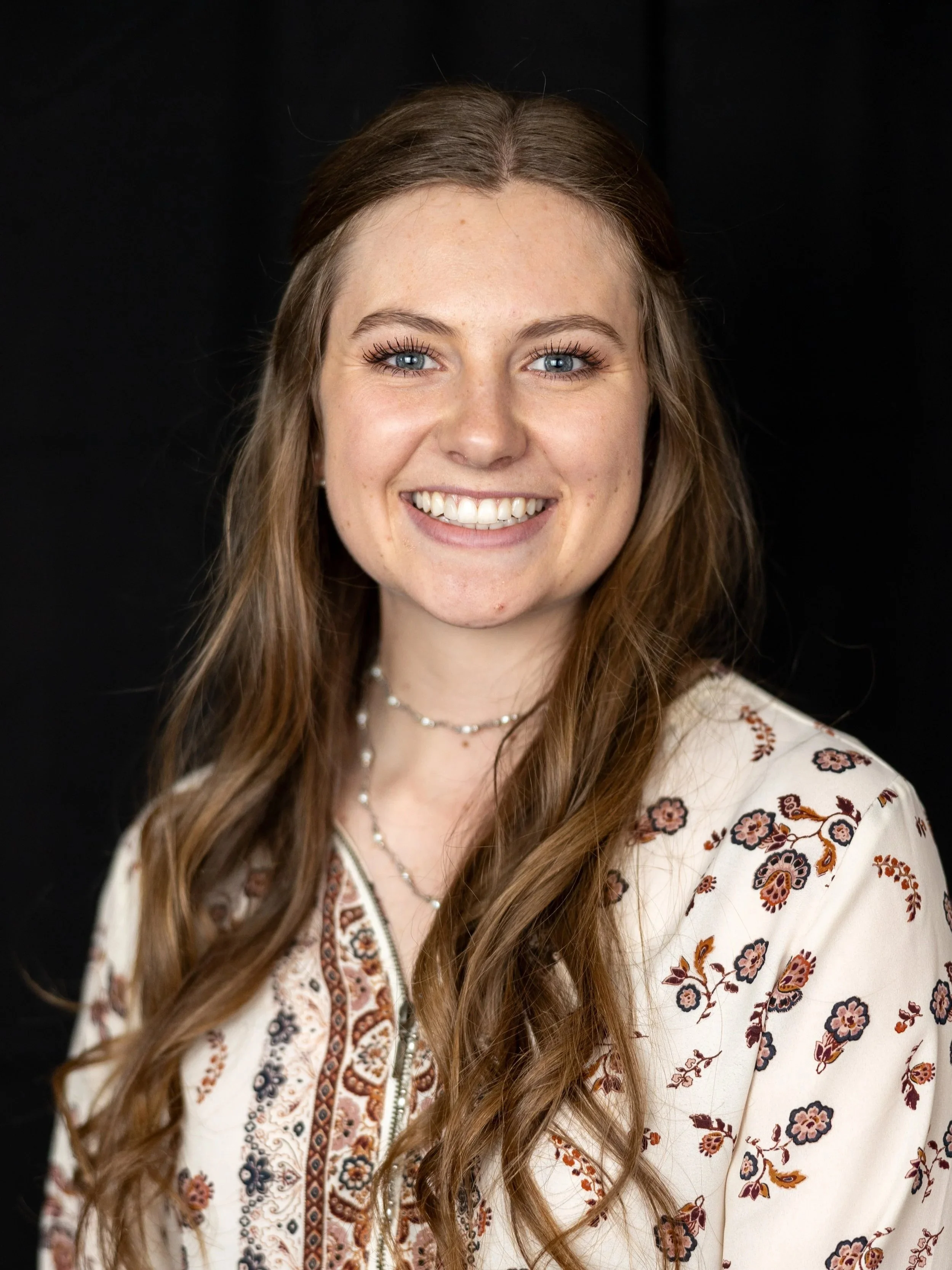 A young woman with long wavy brown hair, blue eyes, and fair skin smiling against a black background, wearing a white floral top and layered necklaces.