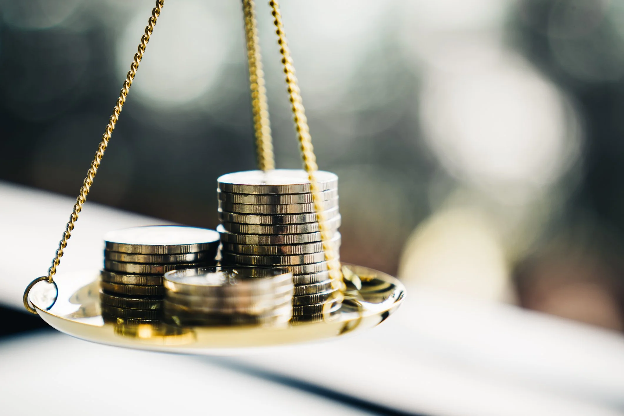 A close-up of a gold-colored balance scale with stacks of silver coins, with a blurred background.