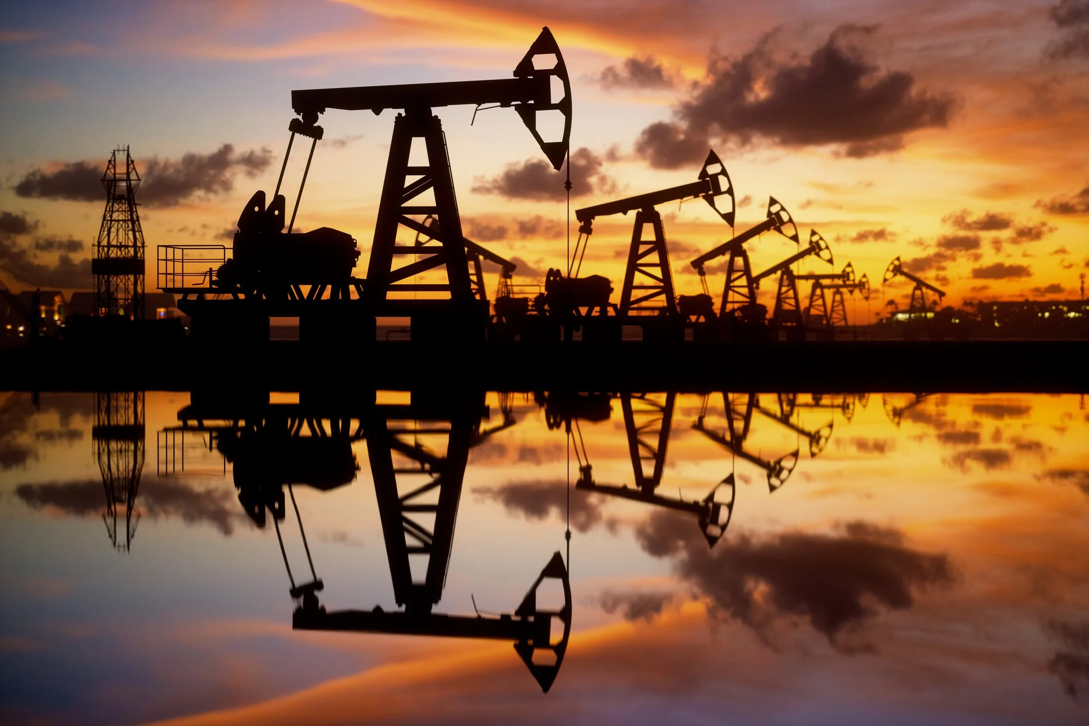 Silhouettes of oil pump jacks against a sunset sky, reflected in water.